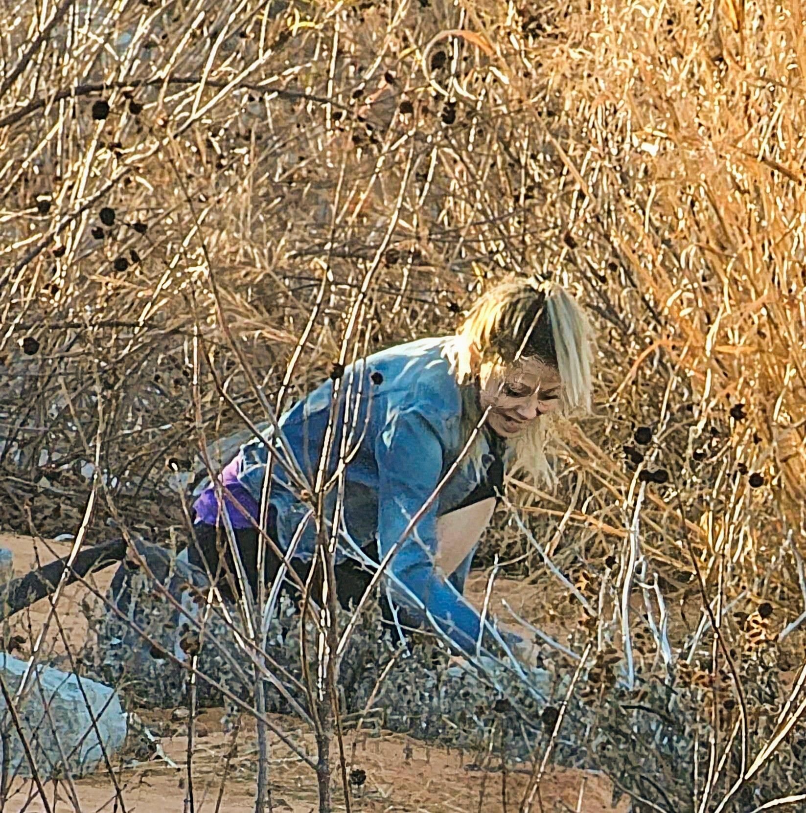 A smiling woman in a denim jacket picking up Rose Rocks out of the sand, surrounded by prairie grass